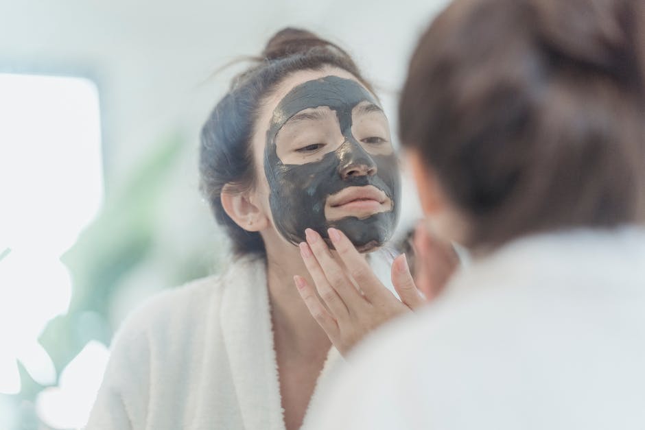 Woman in a robe applies a black facial mask while looking in the bathroom mirror.