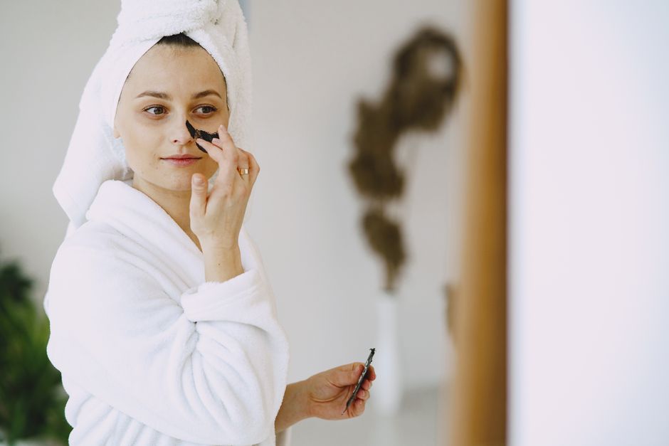 Thoughtful female in white bathrobe with towel on head applying clay mask on face while standing in living room and looking away