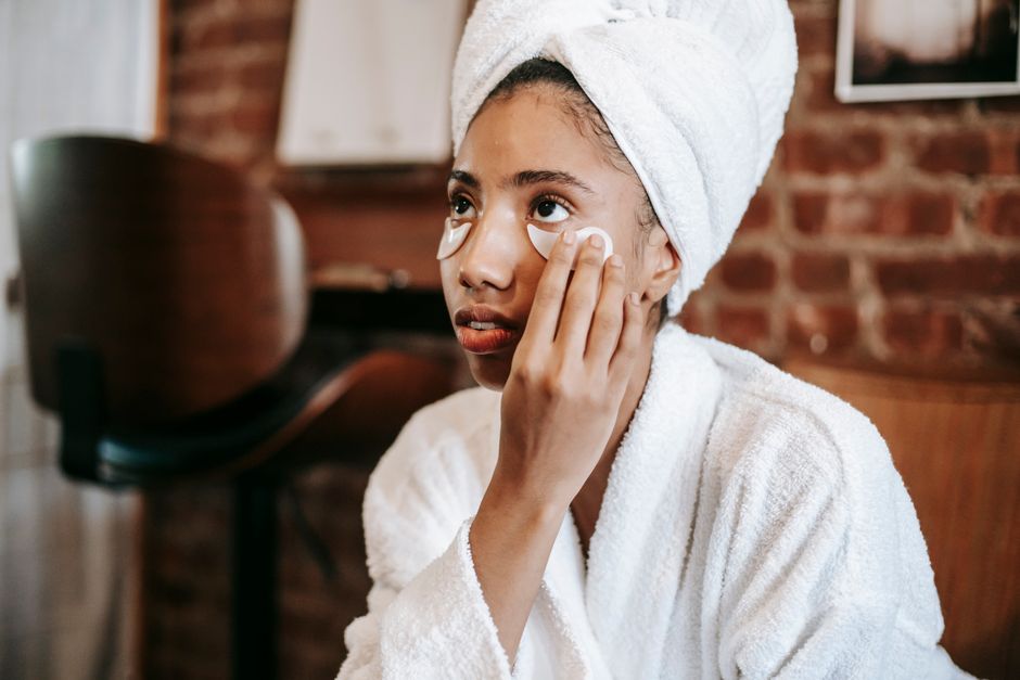 Young African American female with towel on head applying eye patches on face while looking away