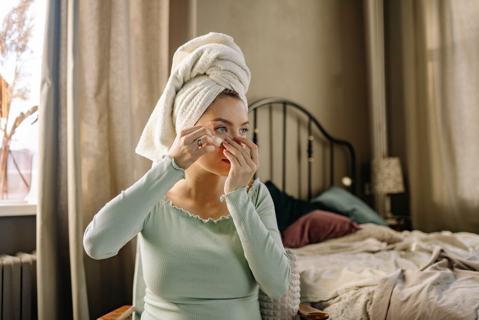 A woman applies beauty products while practicing self care in a cozy, well-lit bedroom.