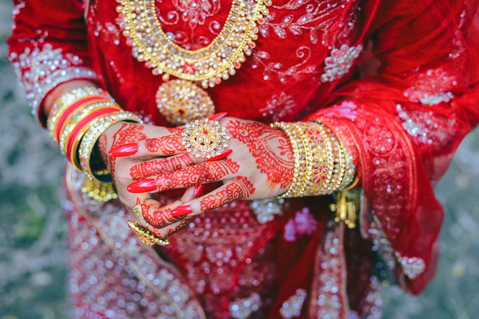 A close-up of a bride's hands adorned with red henna, gold jewelry, and a red dress, showcasing cultural elegance.