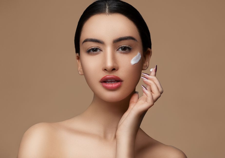 Crop sensitive female with makeup and red lips applying cream on delicate face skin while looking at camera on beige background