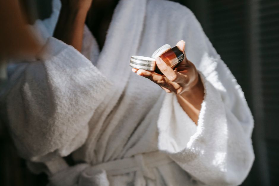 Crop anonymous ethnic female in bathrobe applying moisturizing cream on face while reflecting in mirror in house