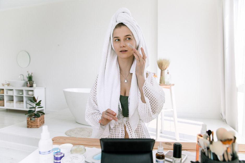 Woman in towel applying face cream in a sunlit bathroom, enhancing her beauty routine.