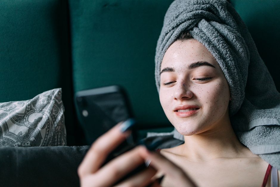 Young woman enjoying skincare routine, relaxed at home with a towel on her head, holding a smartphone.