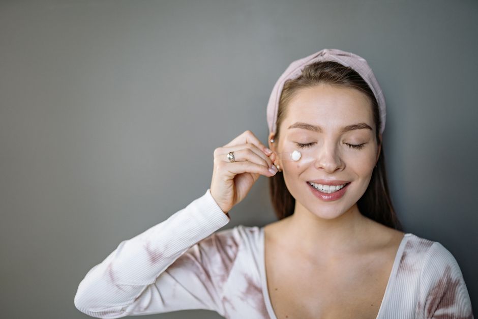 Smiling woman applying skincare cream with eyes closed on gray background.