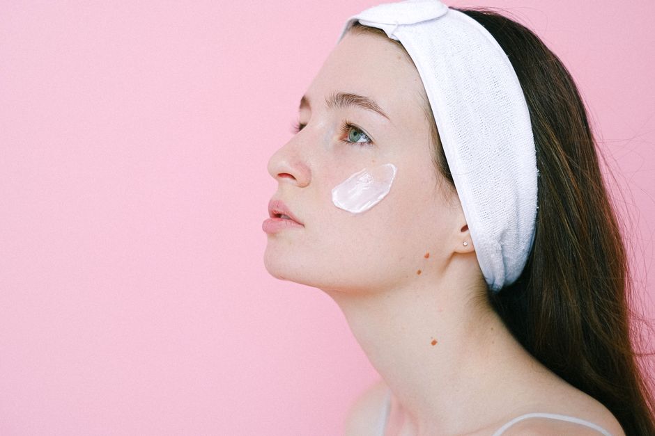 Side view of pensive female wearing headband while standing on pink background with cosmetic product on cheek and looking up