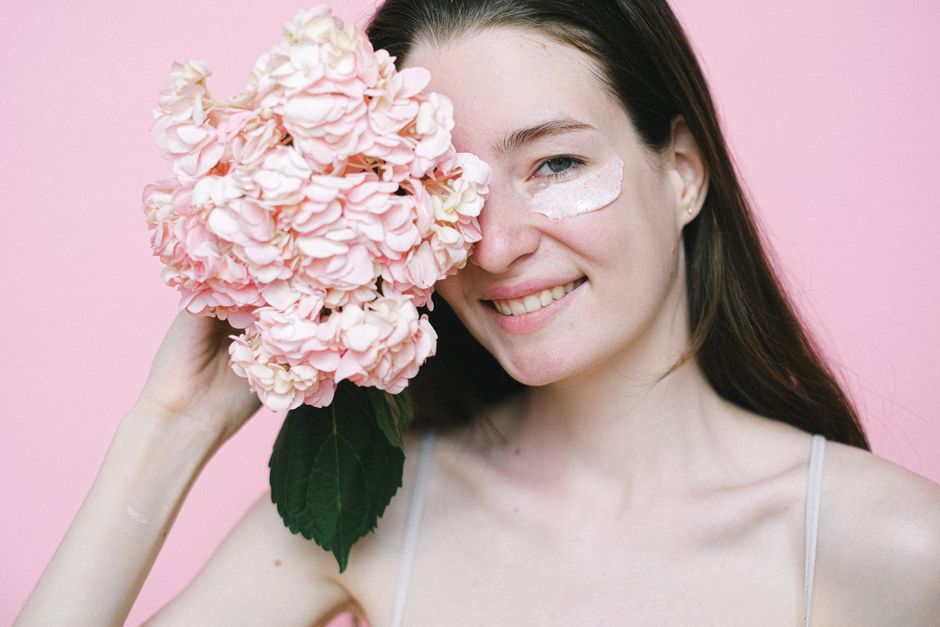 Crop young woman hiding eye behind pink hydrangea while standing with moisturizing  product on face skin against pink background