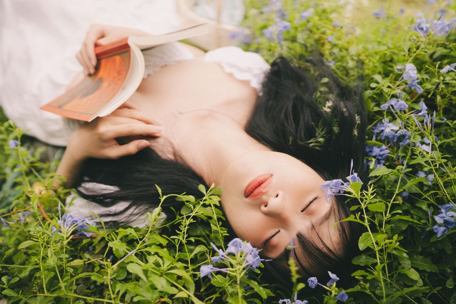 Woman resting with closed eyes and book among purple flowers outdoors.
