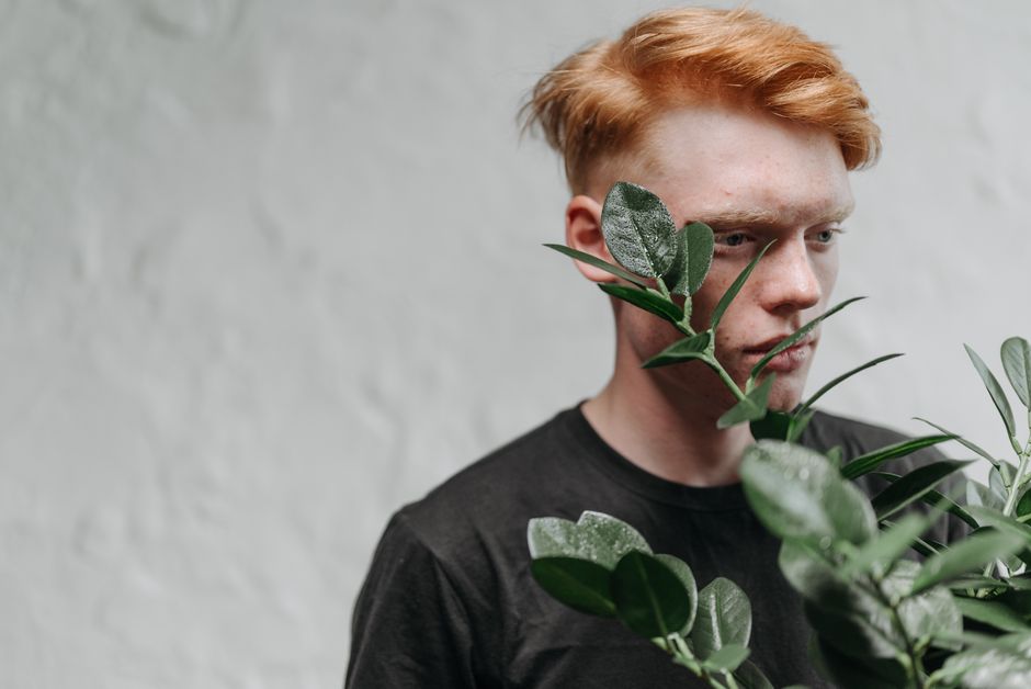 Close-up portrait of a red-haired man in casual attire, partially obscured by green leaves indoors.