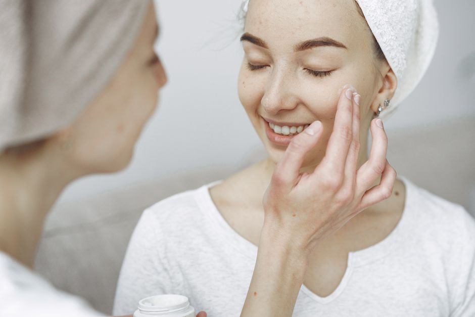 Two women enjoying a skincare routine, applying cream and bonding indoors.