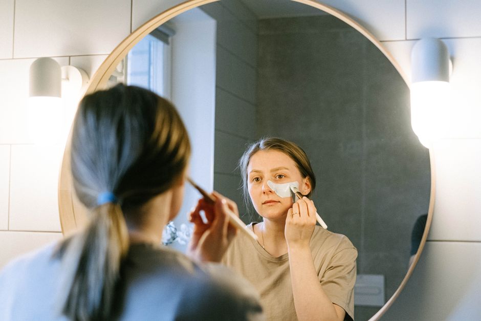 A woman applies facial cream in front of a round bathroom mirror, illustrating a beauty routine.