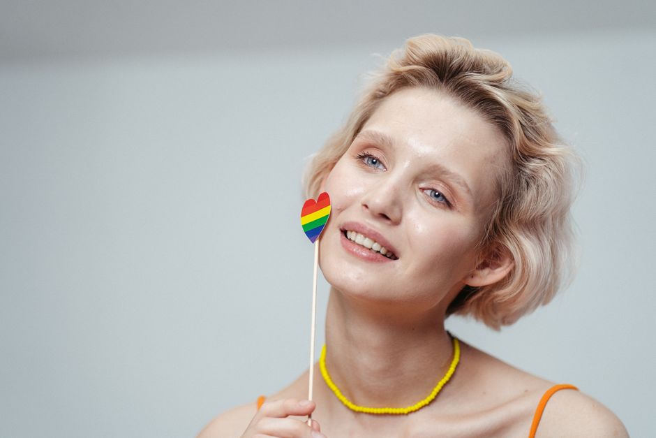 A smiling woman indoors holding a colorful rainbow heart symbol, portraying joy and pride.