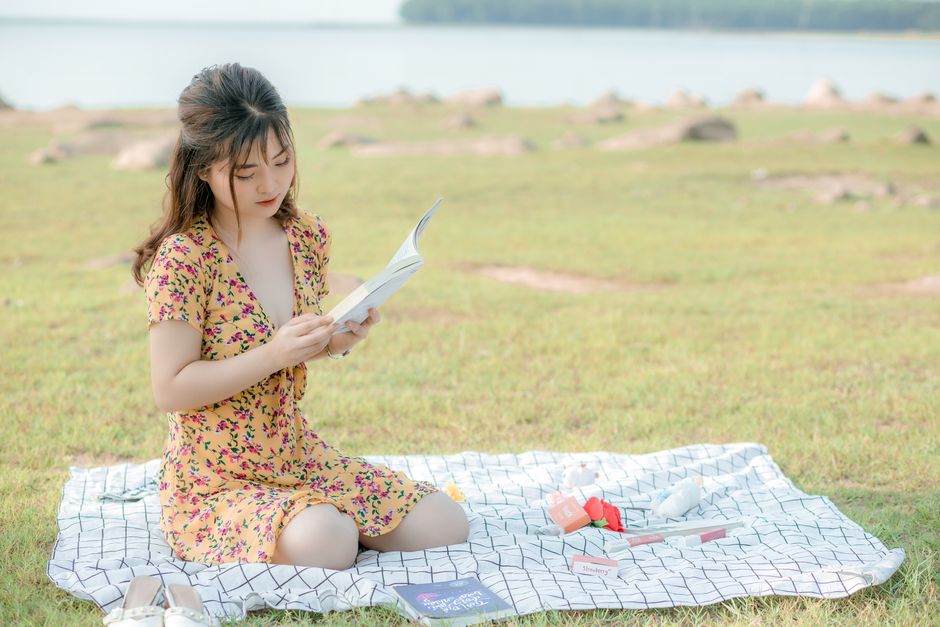 Romantic young Asian female student in light dress sitting on blanket on grassy ground near sea and reading interesting book
