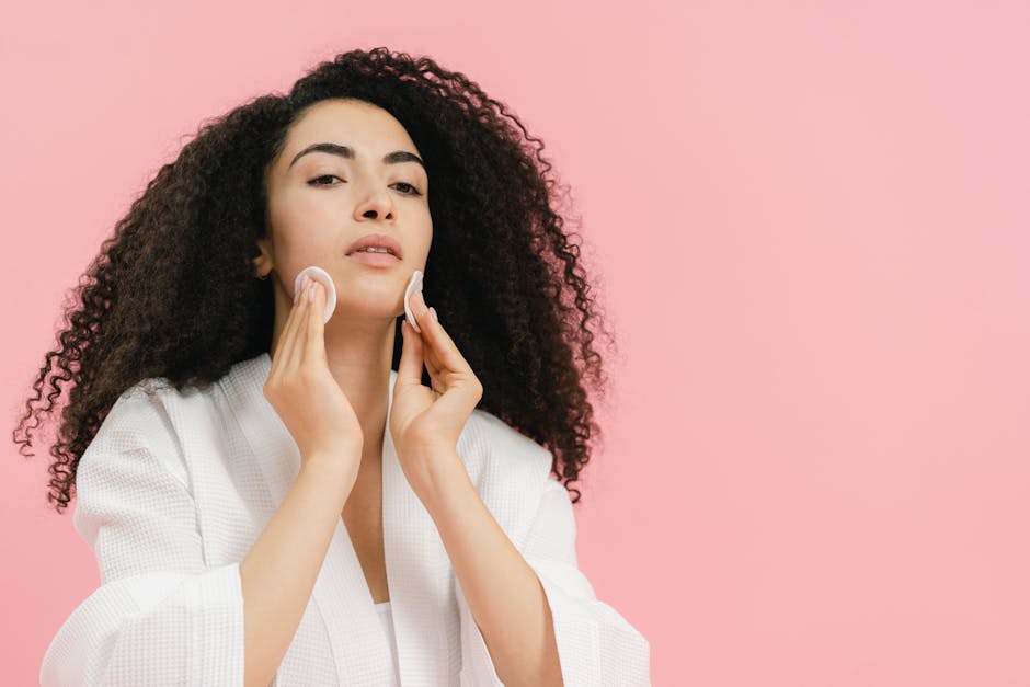 A young woman with curly hair using cotton pads for her skincare routine against a pink background.