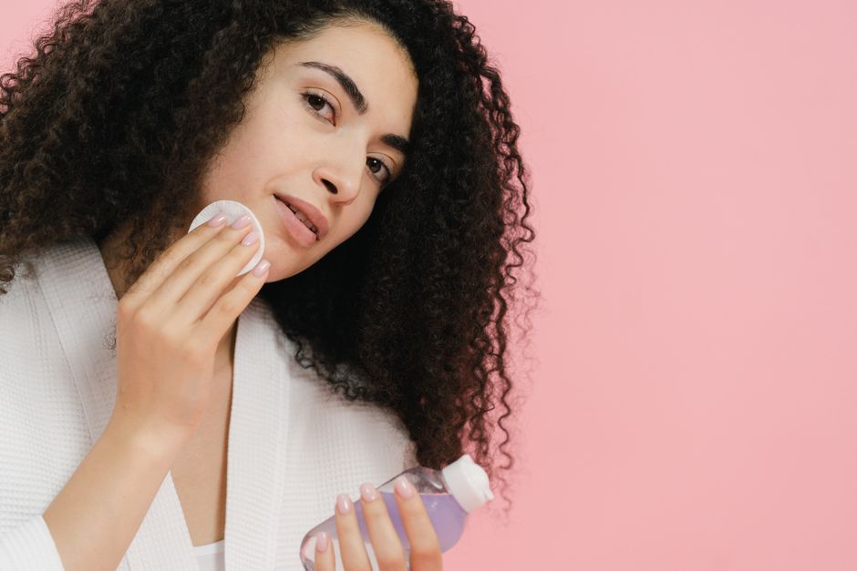 Young woman using a cosmetic pad and product for skincare against a pink backdrop.