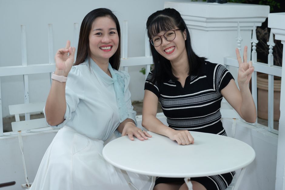 Two cheerful women sitting indoors, sharing a joyful moment with peace signs.