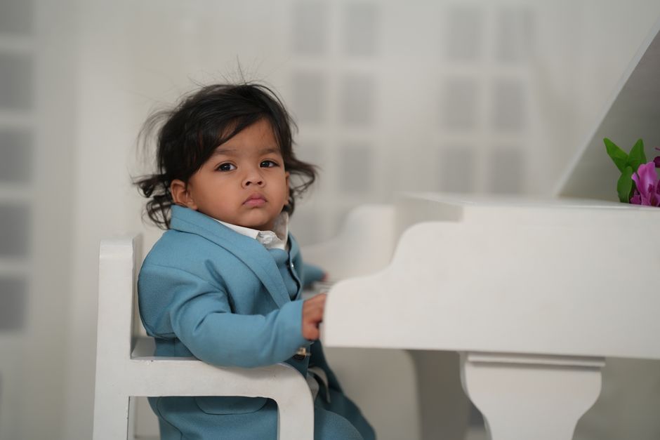 Charming baby in a blue suit sitting at a white piano with pink flowers, indoors.