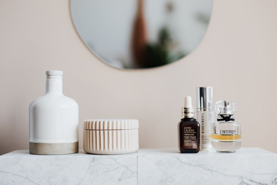 Glass flasks with cream and lotion near perfume and big ceramic vase on marble surface close to beige wall with oval shaped mirror