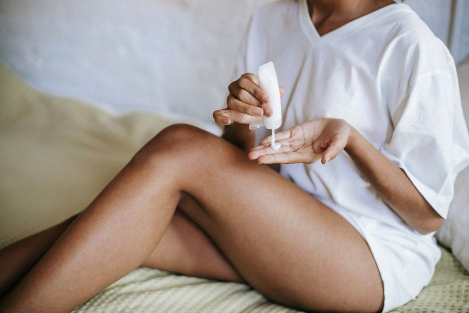 Unrecognizable female in white t shirt with bared legs sitting on bed and applying cream on hand while doing skincare routine