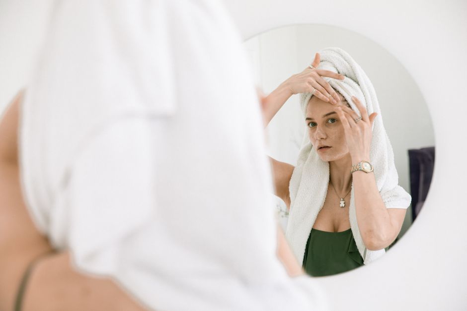 Woman with towel on head applying skincare, enjoying beauty routine in front of mirror.